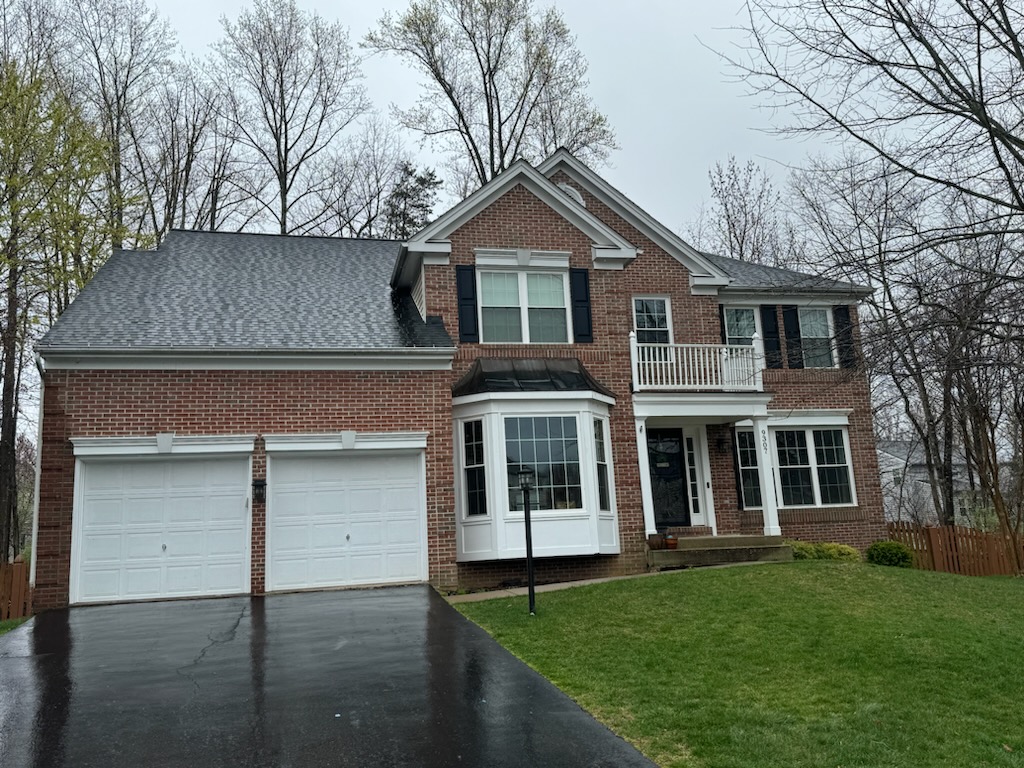 Brick colonial with a first-floor bay window, second-floor balcony, and two-car garage on a rainy day.